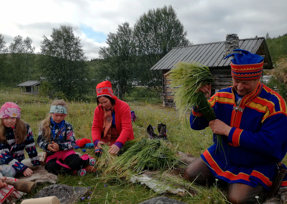 Saamelaisasuihin pukeutuneet mies ja nainen sekä kaksi lasta niputtavat kenkäheiniä.