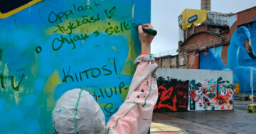 Lapsi valkoisessa maalaushaalarissa kirjoittaa kiitoksia ulkoseinälle, jossa graffiteja.